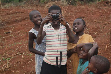 Children in Africa using a camera, smiling, and looking at it outside on reddish dirt.