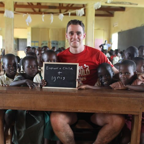 Man in red shirt with a chalkboard, surrounded by Black children in a classroom setting.