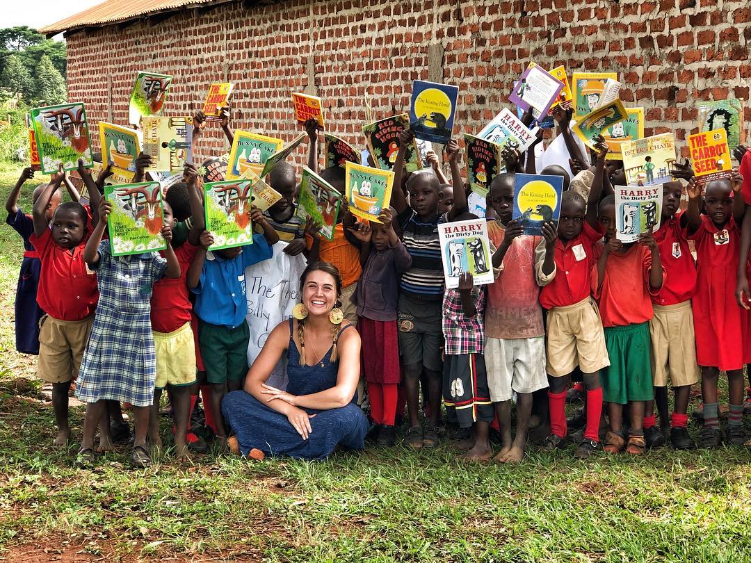 Woman kneels with a group of children holding up books outside a brick building.