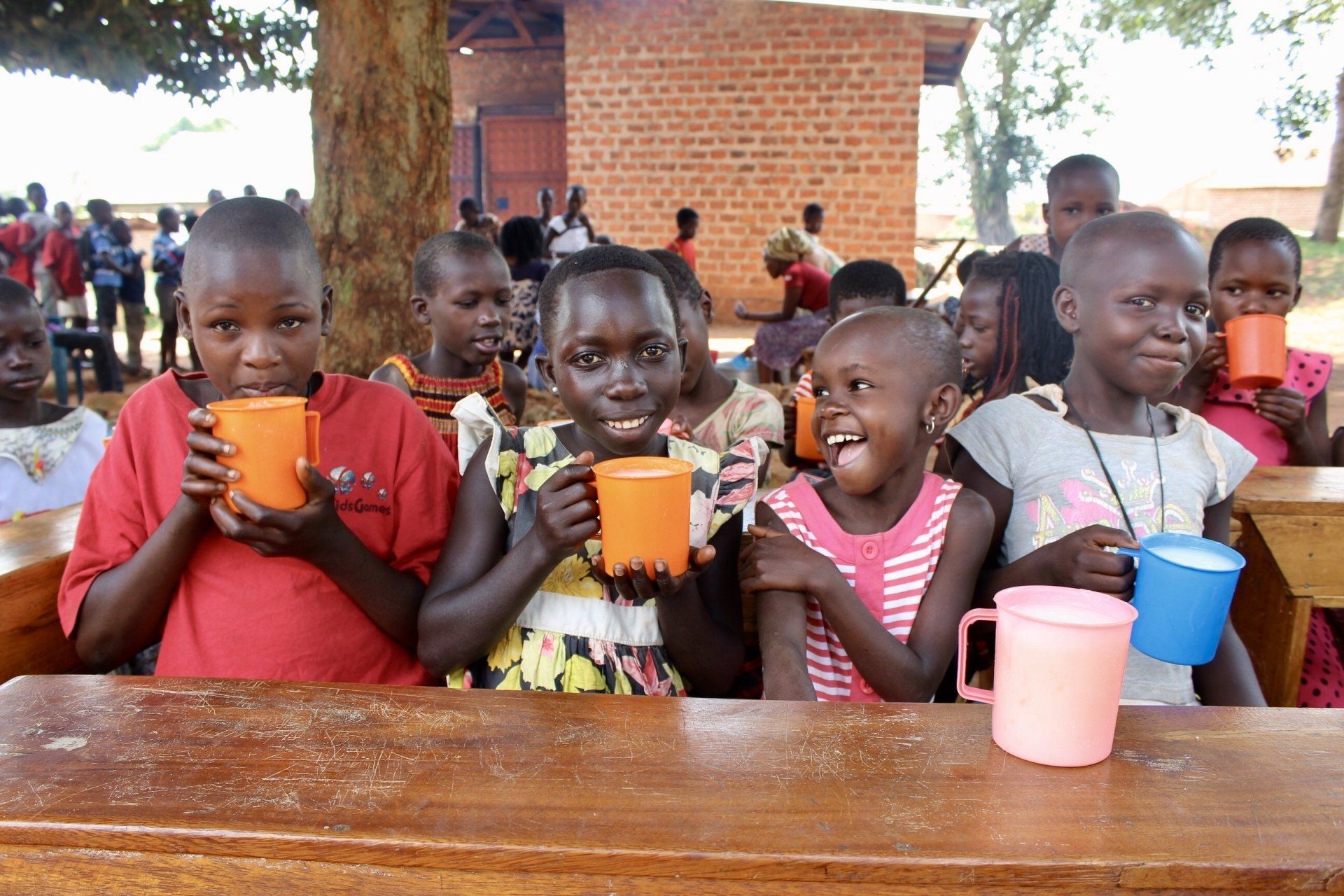 Children at a table drinking from orange and blue cups, smiling outdoors.