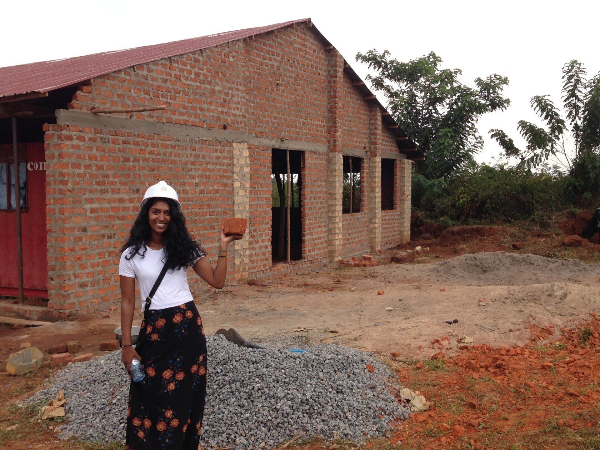 Woman in construction hat holds brick, standing in front of a building under construction in an outdoor setting.