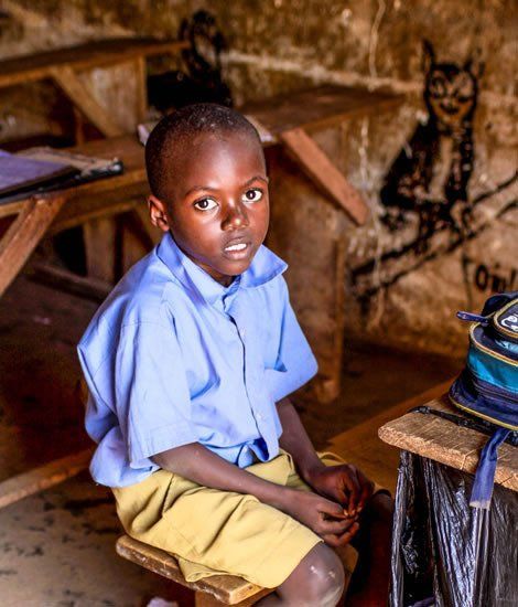 A young boy in school uniform sits in a classroom, looking directly at the viewer.