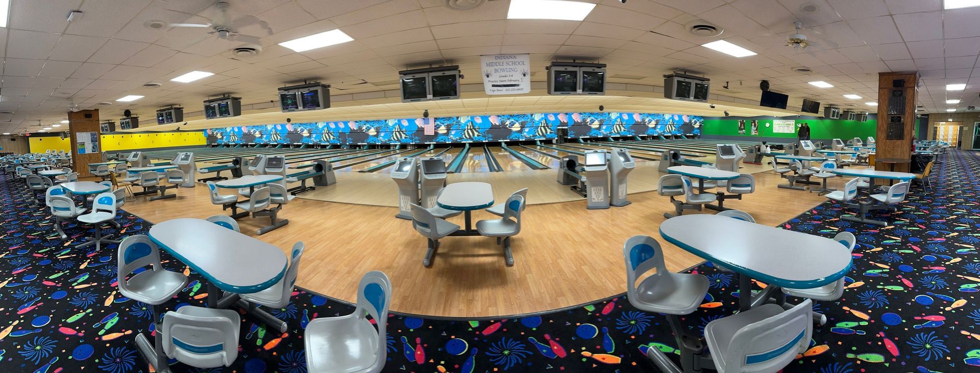 A wide-angle view of a bowling alley with tables, chairs, and bowling lanes with a blue patterned wall in the background.