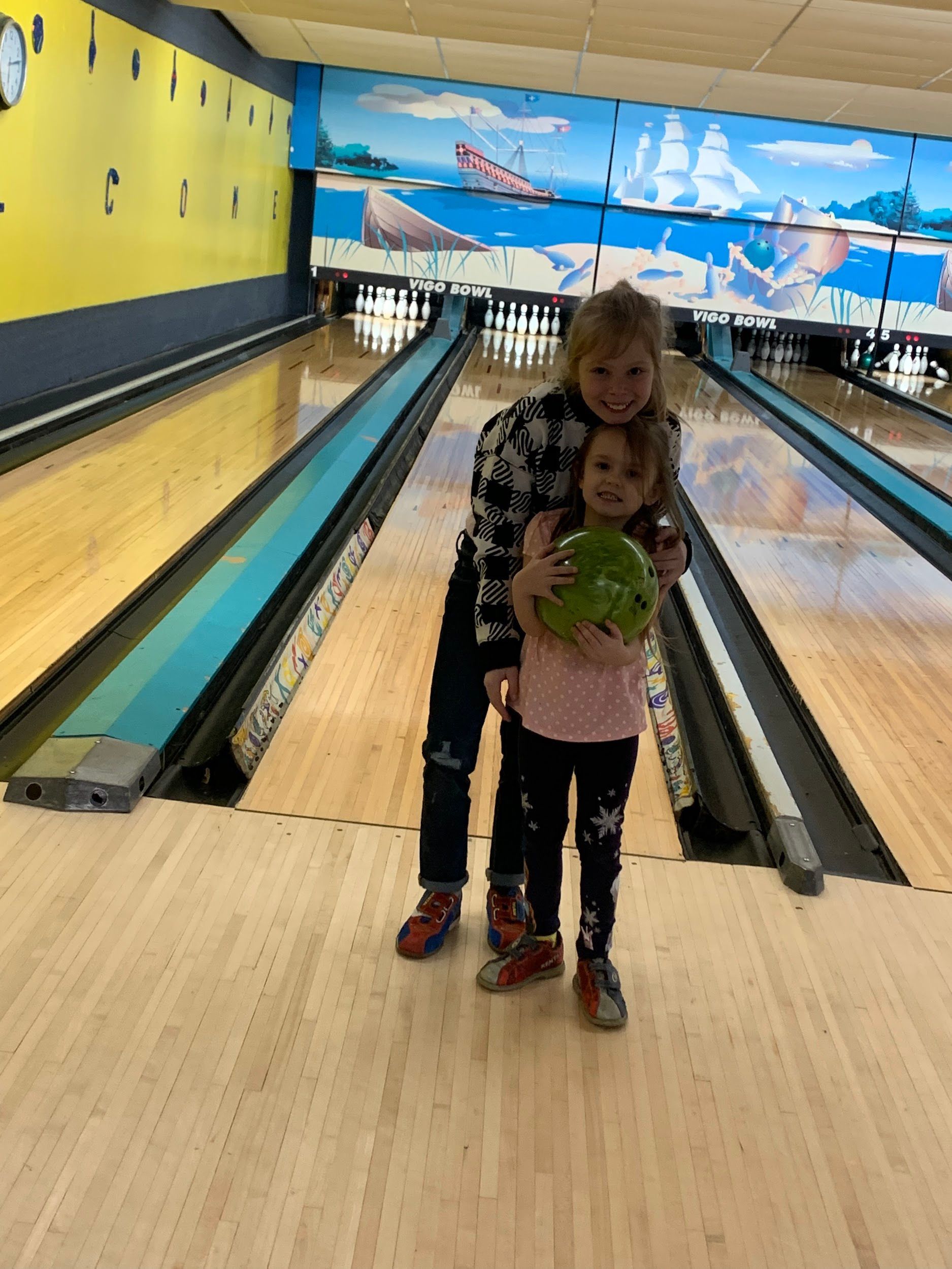Two young girls are standing in a bowling alley holding a bowling ball.
