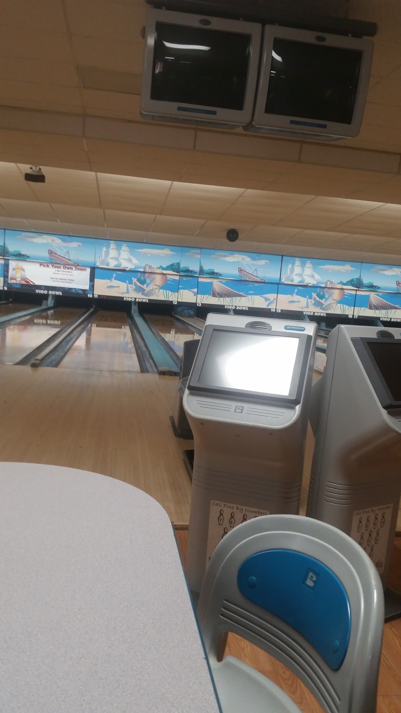 A bowling alley with a table and chairs and two televisions hanging from the ceiling.