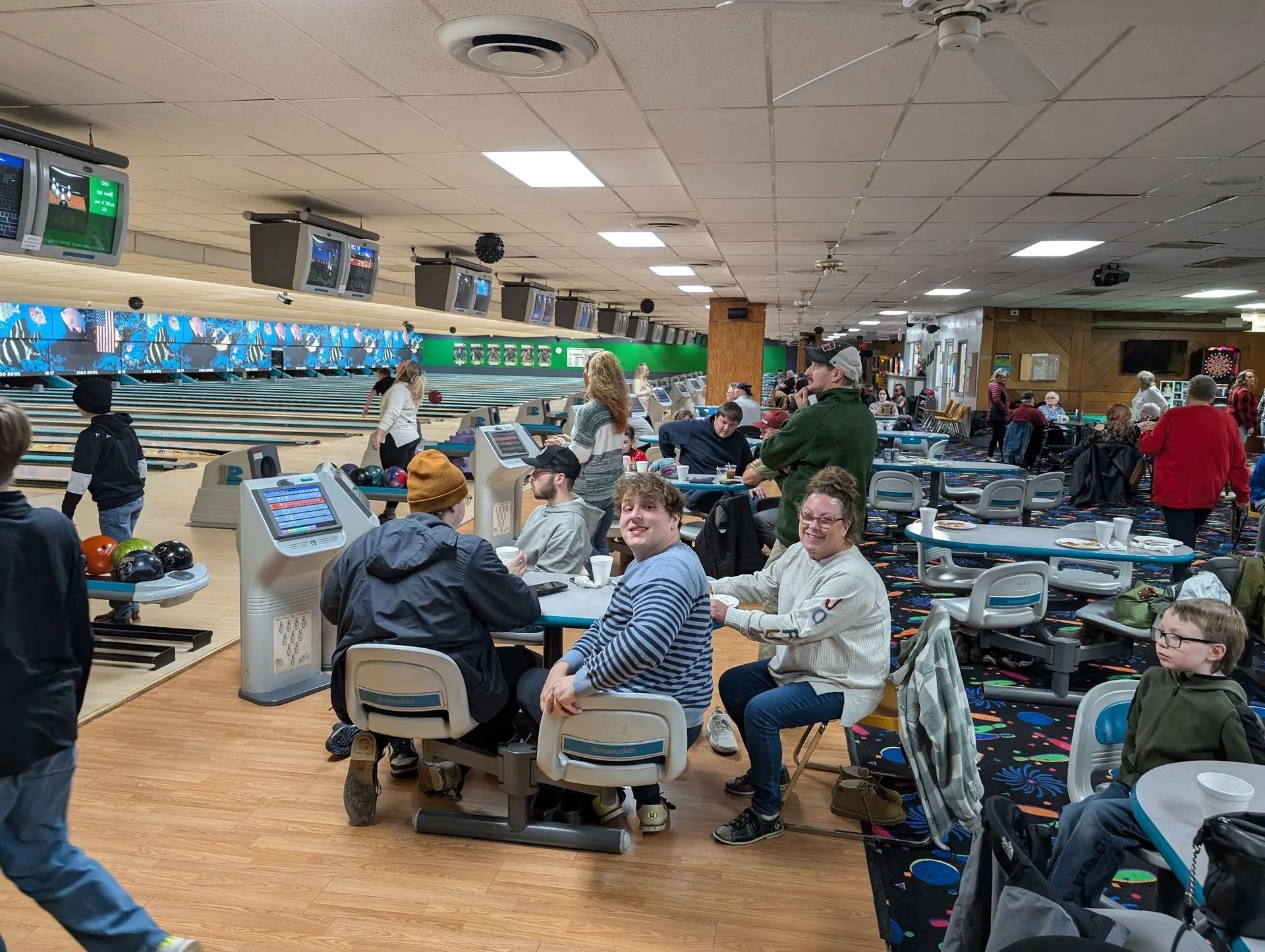 Bowling alley scene: people seated at tables, playing, some smiling, with bowling lanes in the background.