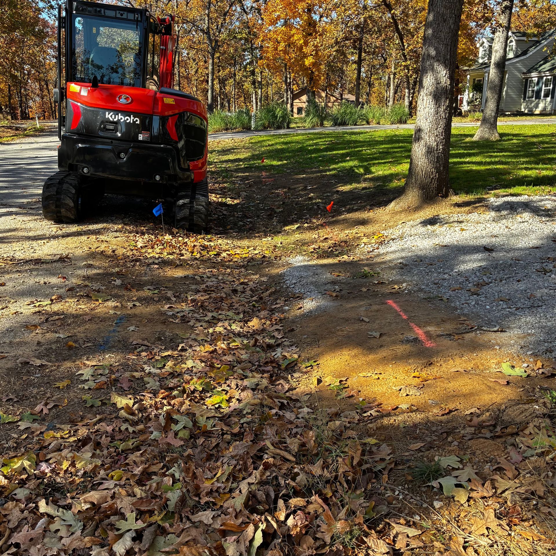 Red Kubota mini excavator on gravel, marked path, fallen leaves, trees, and grass.