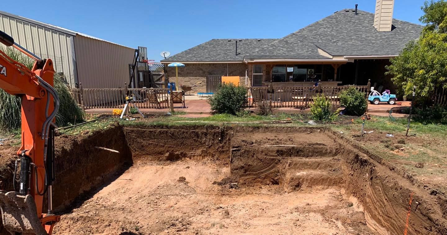 An excavator digs a pool pit in a backyard. The house is in the background.