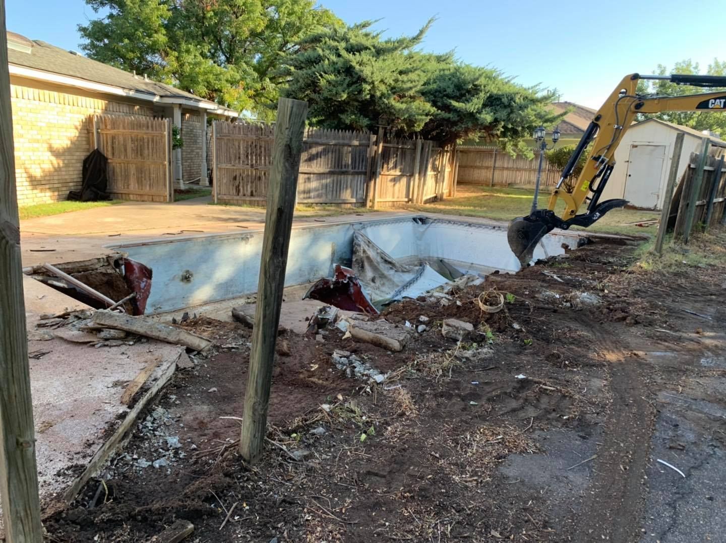 An excavator demolishes a swimming pool in a backyard; the pool is empty and crumbling.
