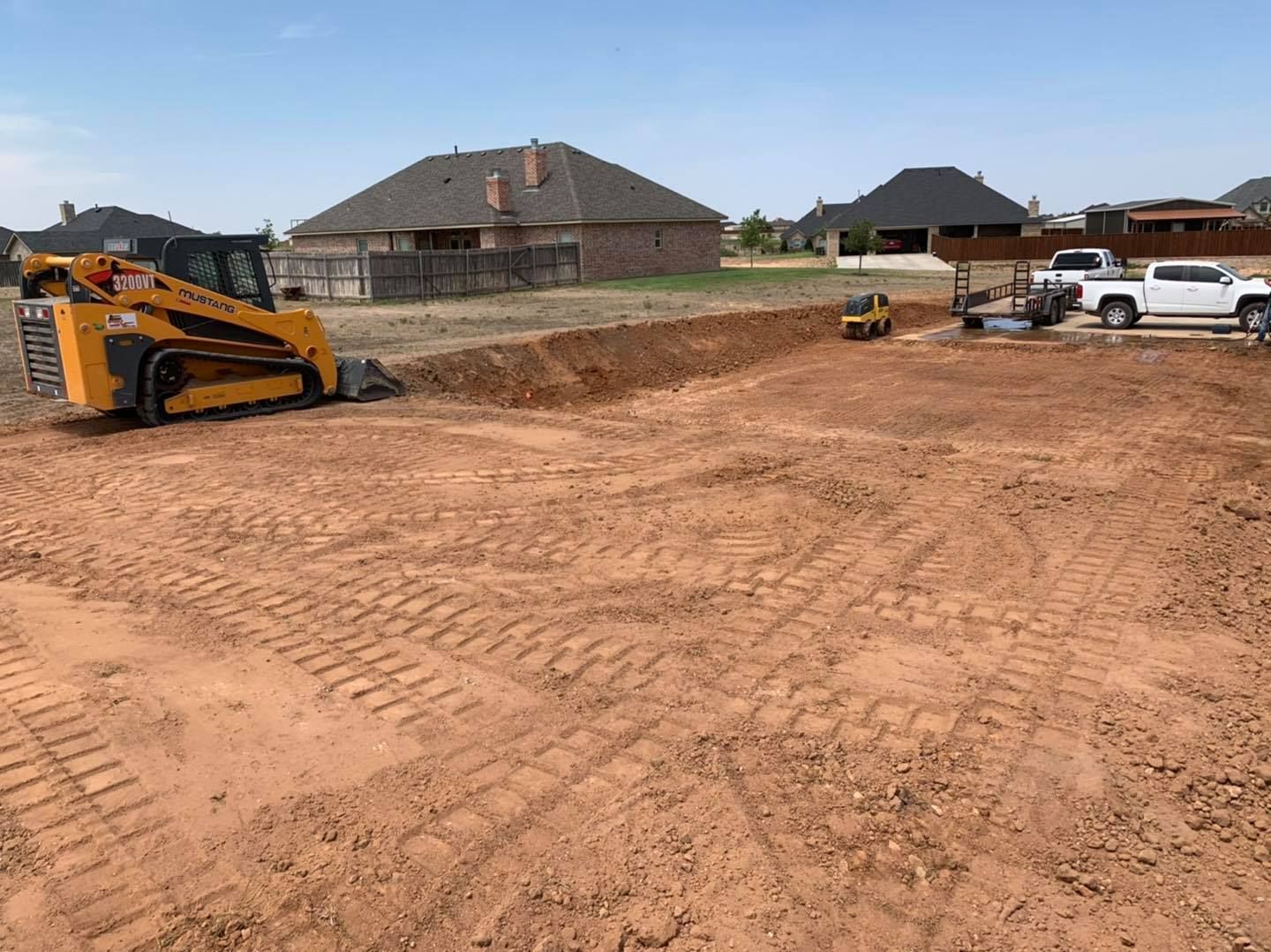 Construction site with skid steer, small excavator, and a pickup truck on red soil, residential area.