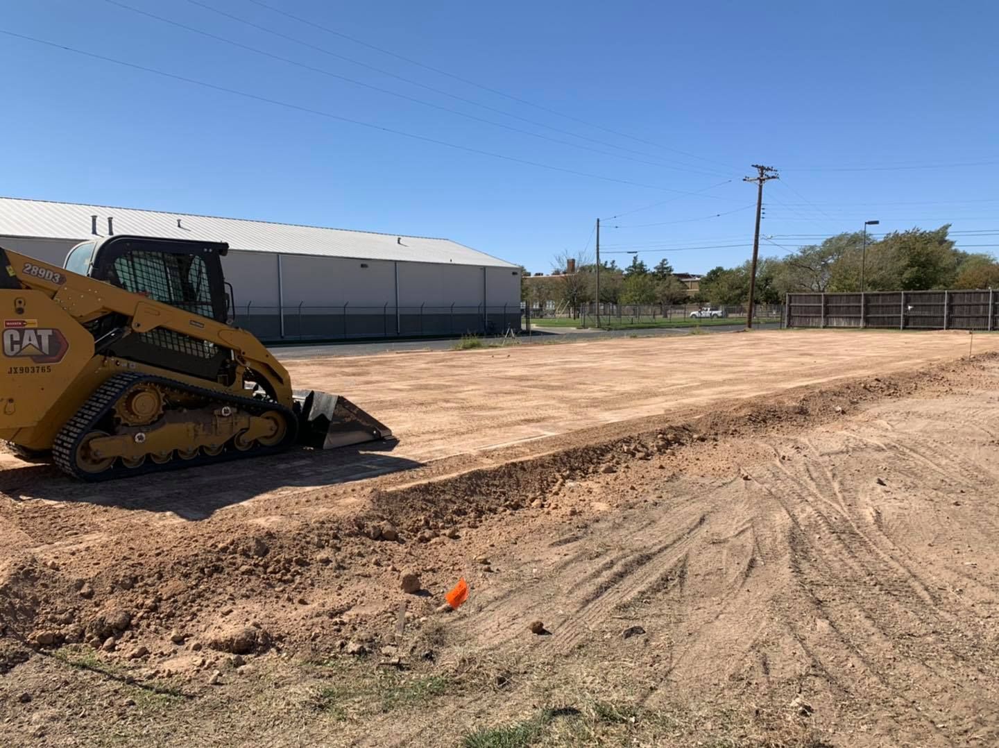 A yellow skid steer tractor digging a trench in a dirt lot under a blue sky.