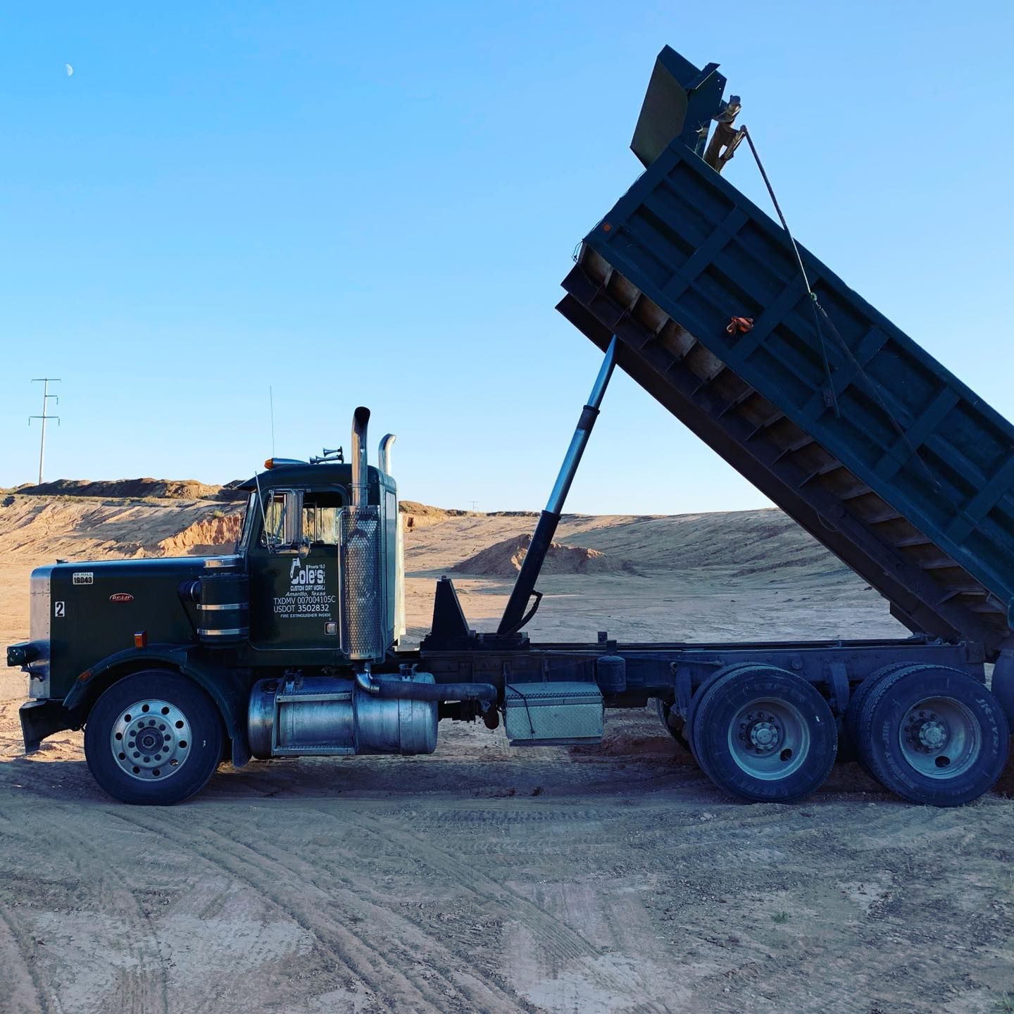 Dark green dump truck with bed raised, against a clear blue sky.