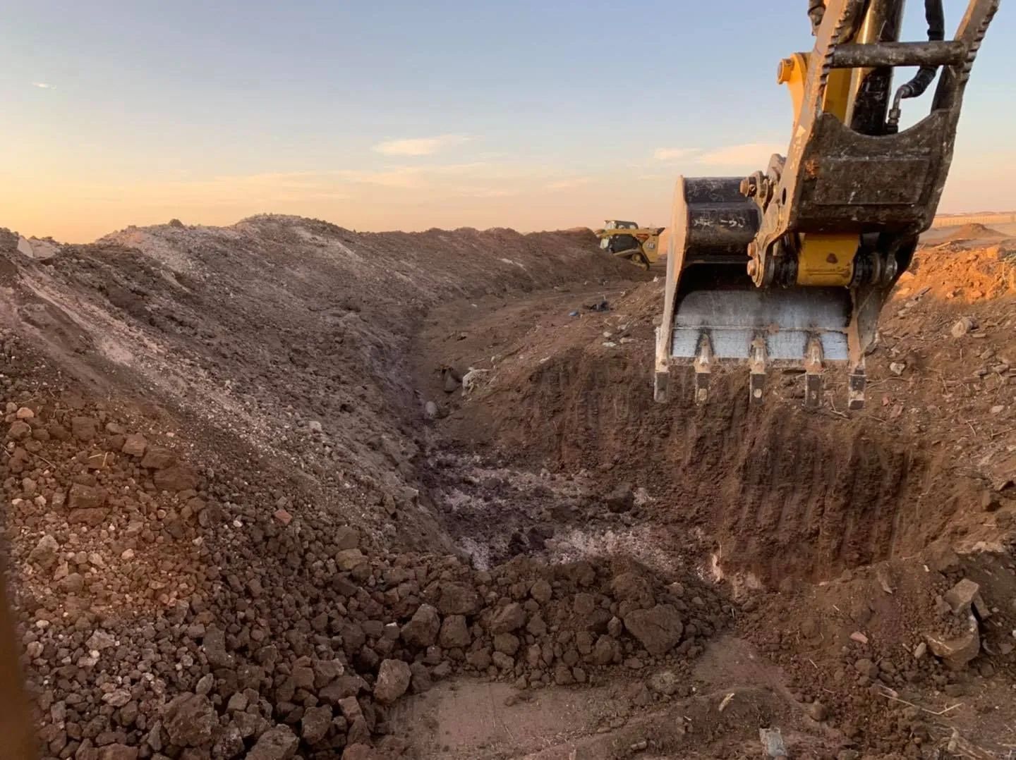 Excavator digging a trench in dirt at a construction site with a sunset in the background.