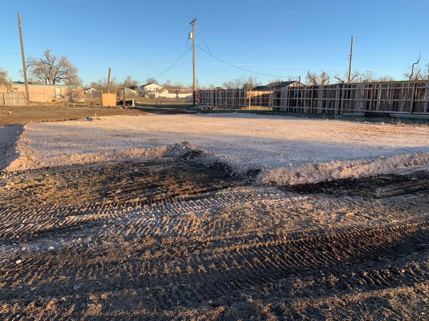 A cleared lot with gravel and dirt, wooden fences, and distant houses under a blue sky.