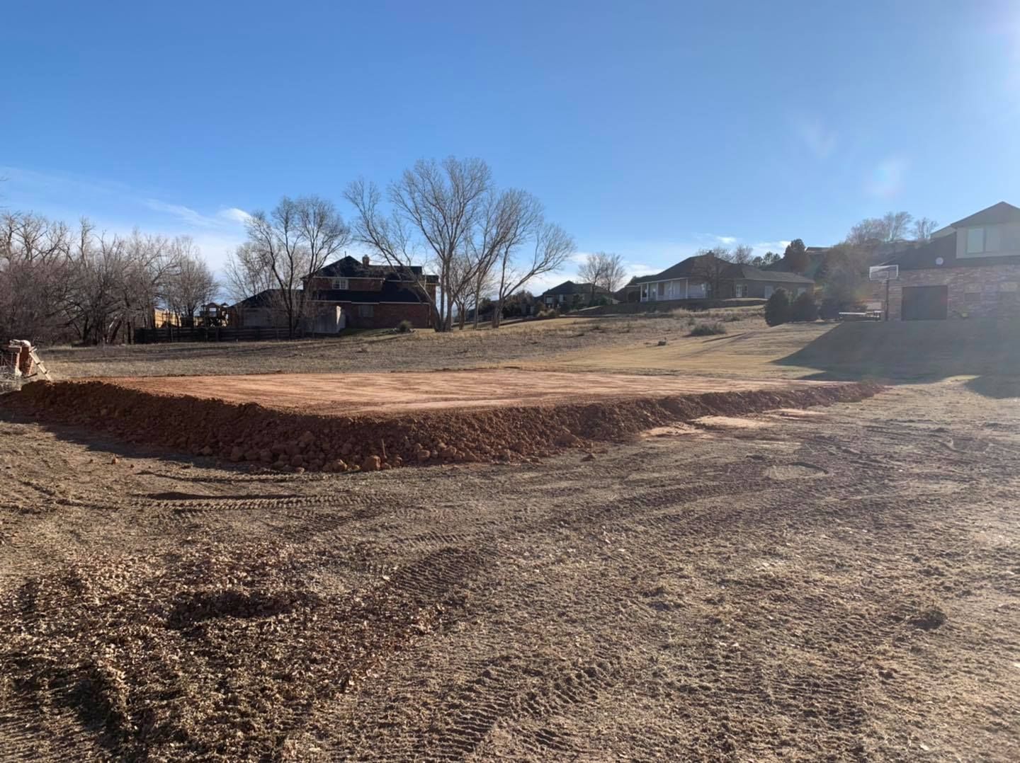 A dirt lot, some brown soil, a pile of reddish gravel, homes, and trees under a blue sky.