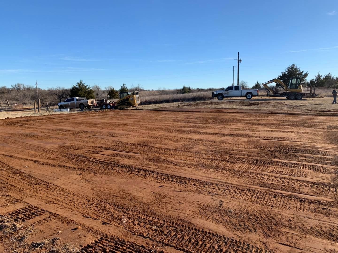 Red dirt field with tire tracks, trucks, and heavy machinery under a blue sky.