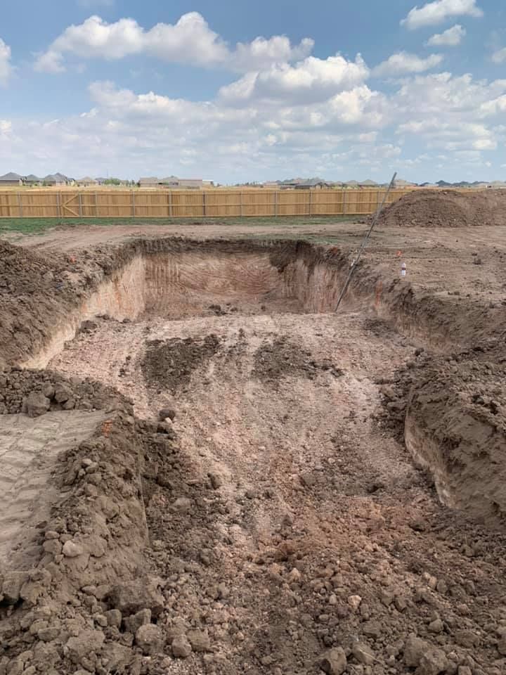 Excavated square foundation pit in a dirt lot, ready for construction. Brown soil under a cloudy sky.