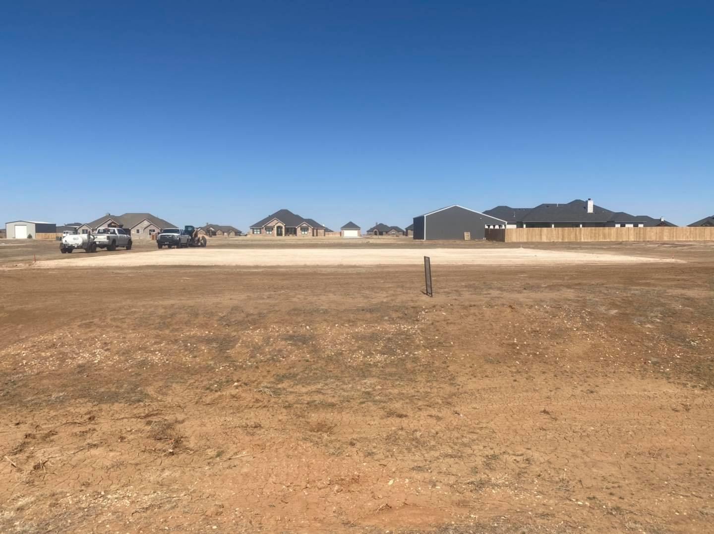 Brown field with a cleared area, partially constructed houses in background, clear blue sky.