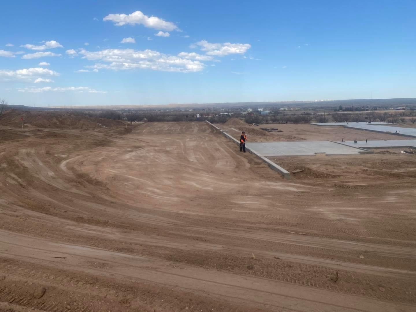 Dirt road with a person on a motorcycle, with a cityscape in the background under a blue sky.