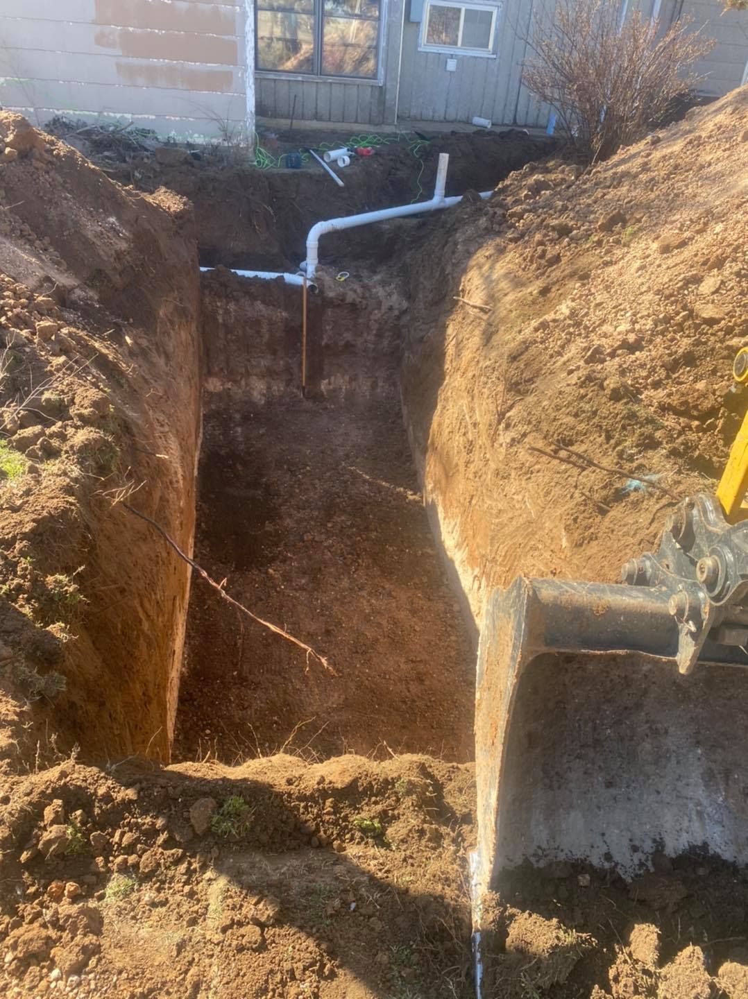 A deep trench dug in brown earth with white pipes visible. A small excavator is at the right side.