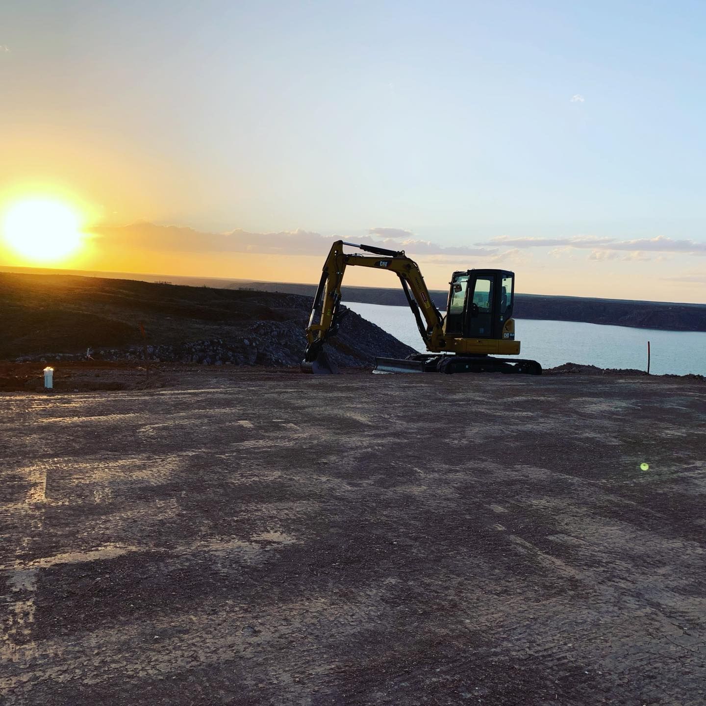 Excavator on a rocky area with a lake and sunset in the background.