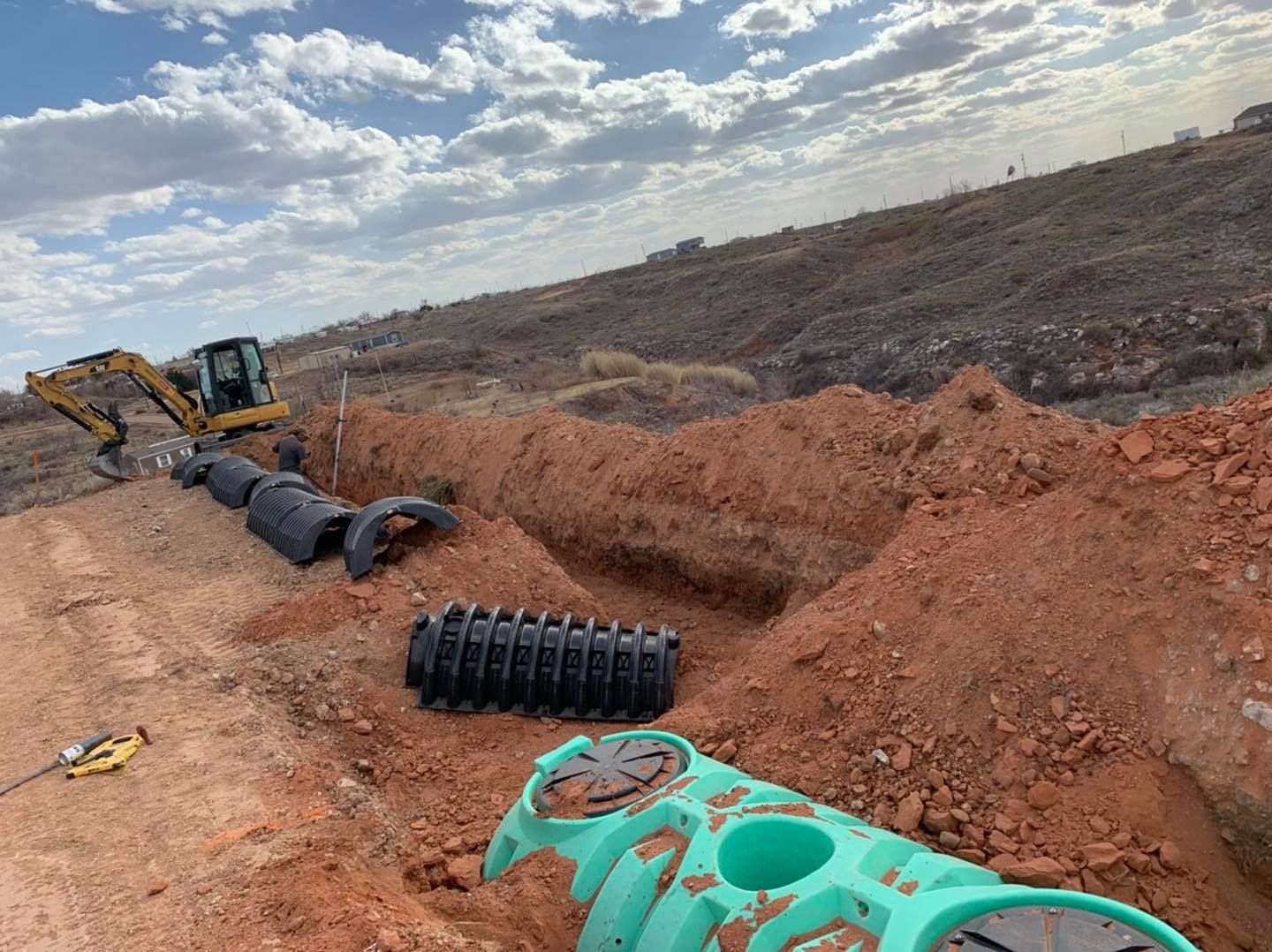 Construction site with excavator, pipes in a trench, and a dirt hillside under a cloudy sky.