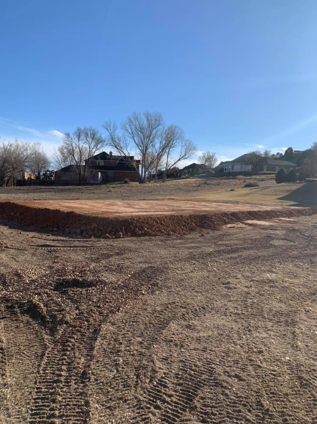 Dirt field with a berm, houses and trees in the background on a sunny day.