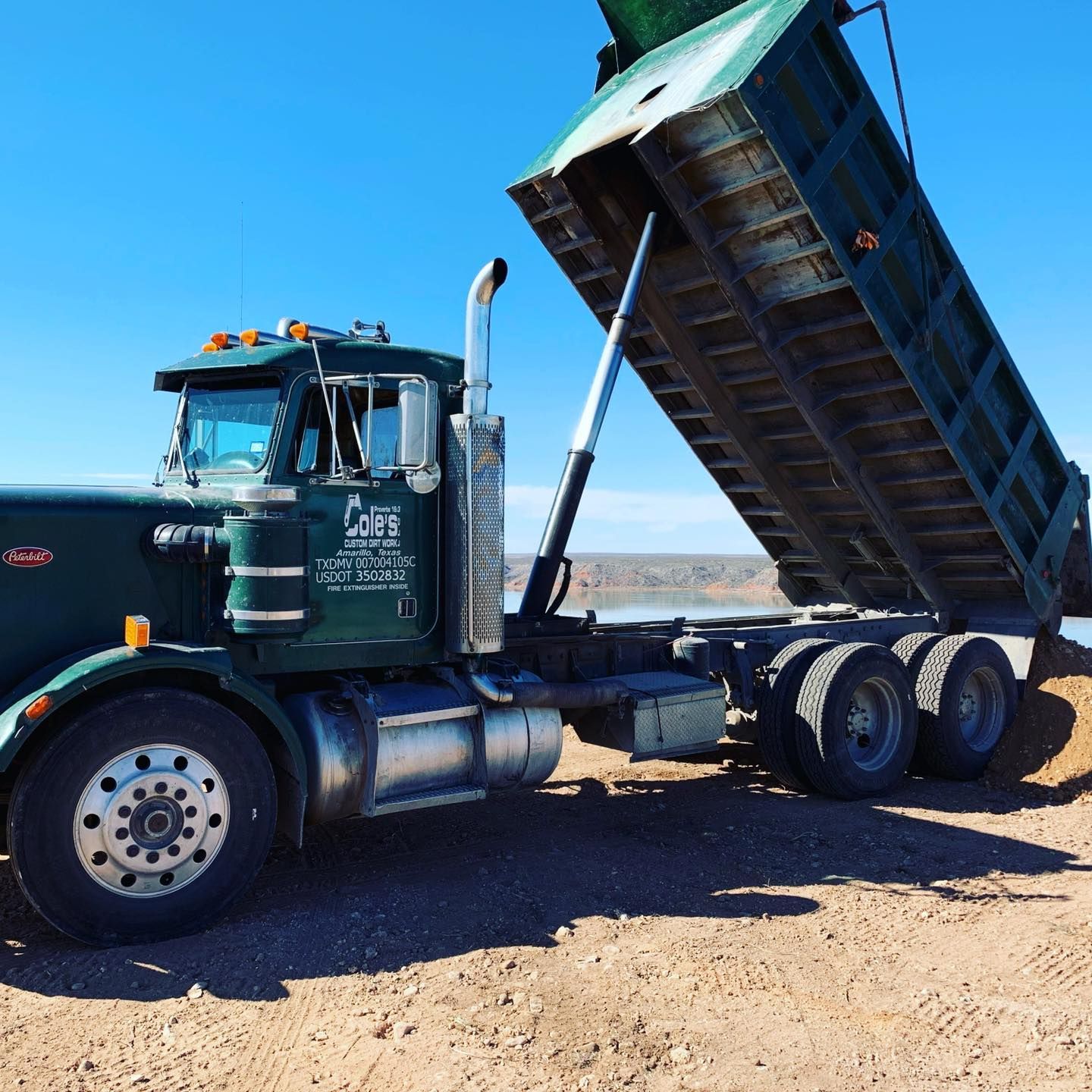Green dump truck with its bed raised, dumping a load of material in a desert setting.