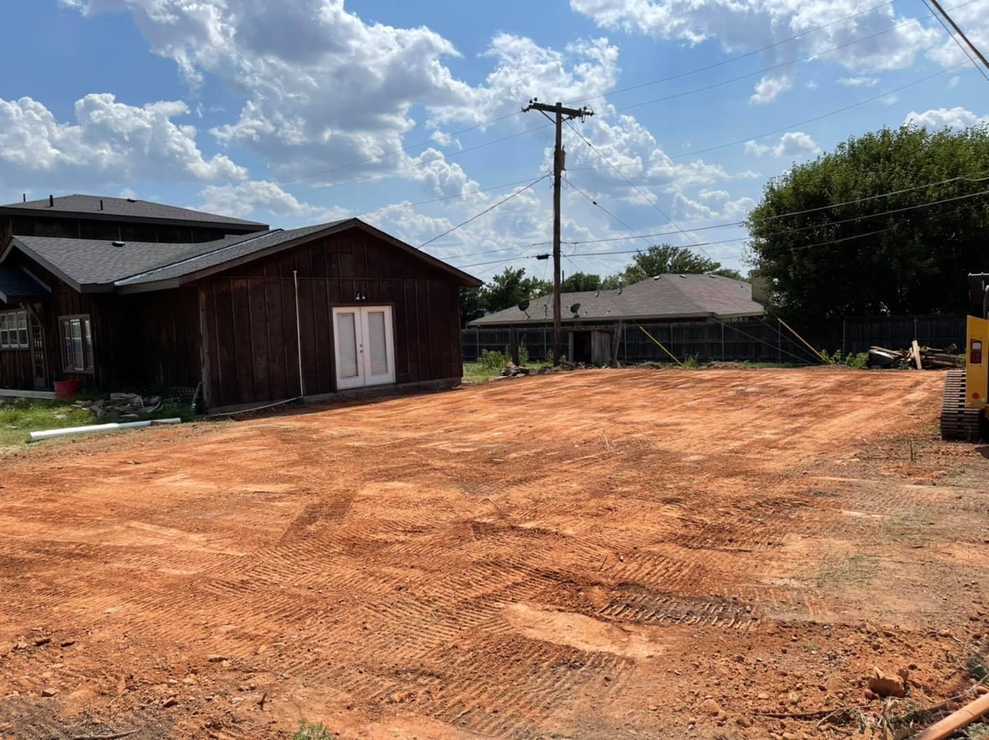 Cleared, red-dirt lot with a small, weathered building and a house in the background, under a cloudy sky.