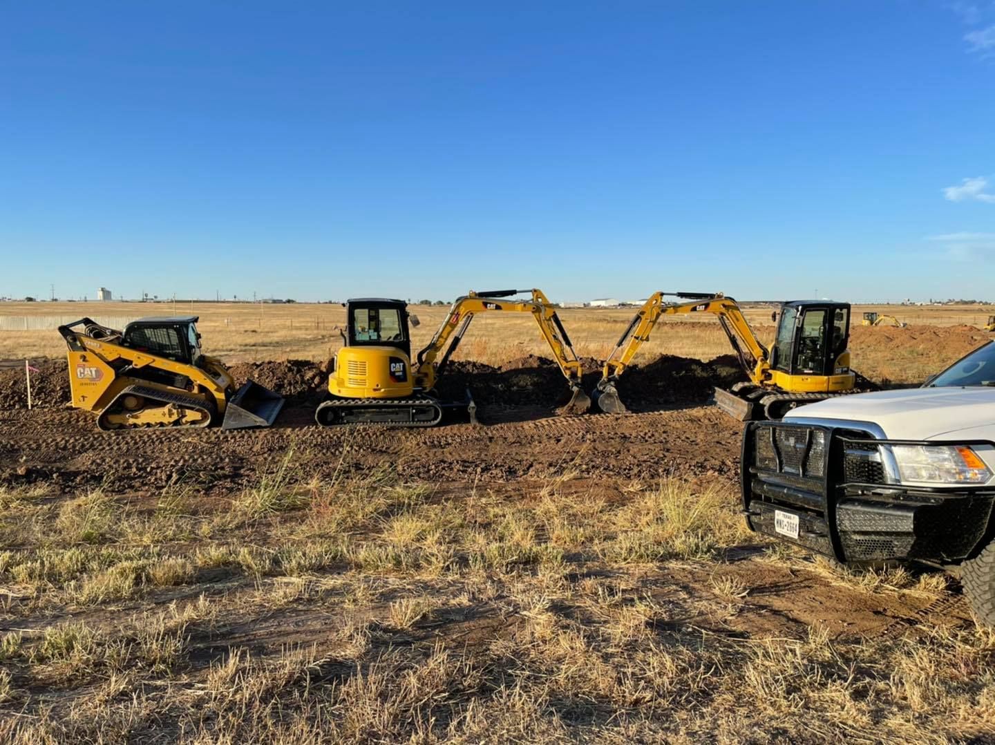 Four yellow construction vehicles digging in a dirt field under a blue sky.