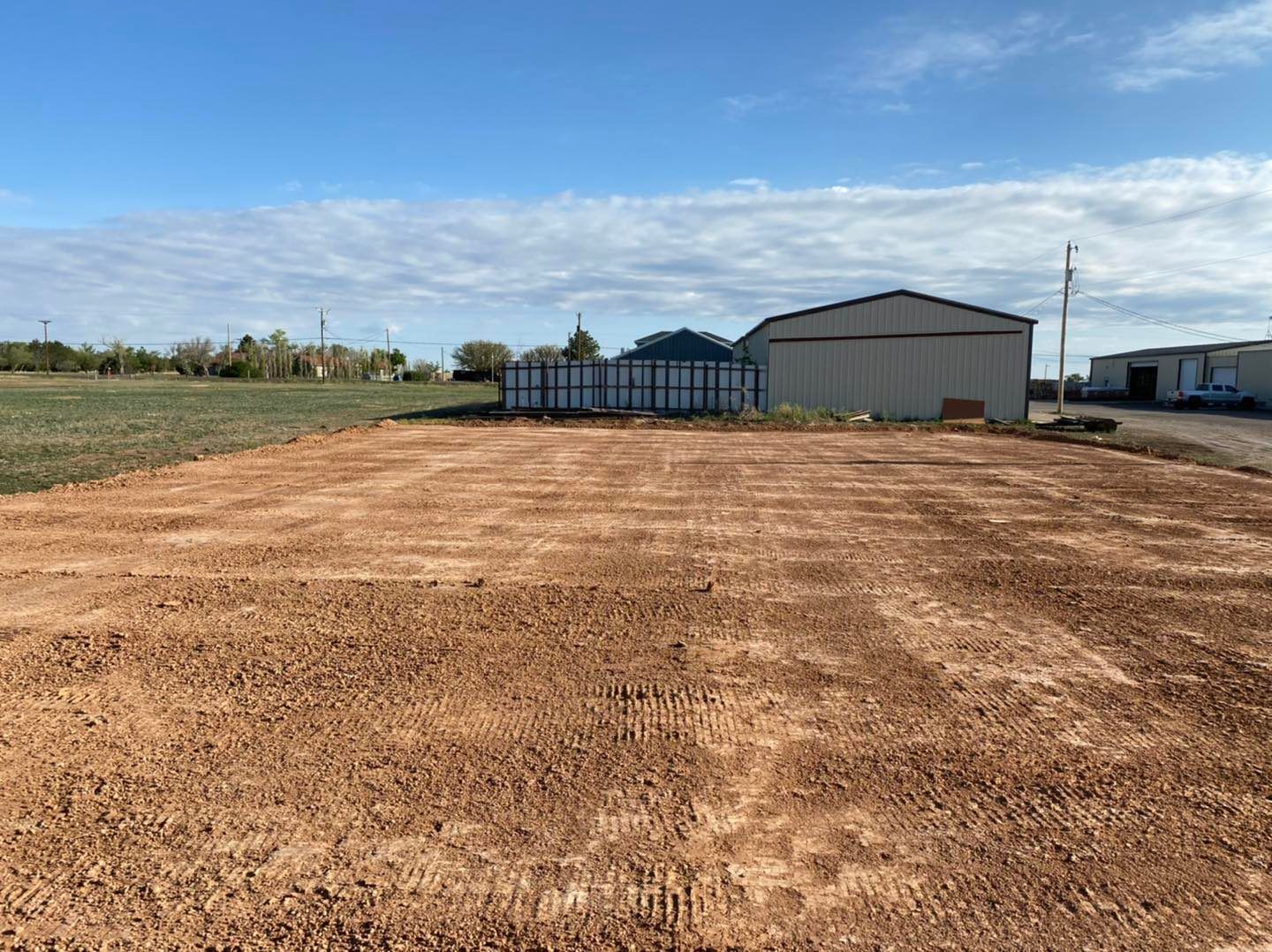 Flat, red-brown earth in foreground; a metal barn and containers in background, under blue sky.