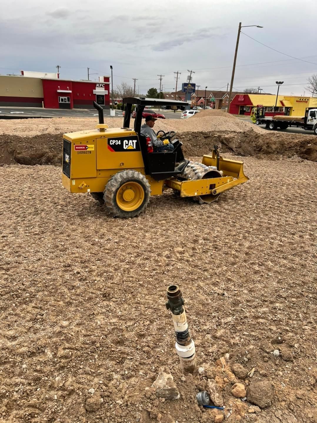 Yellow Caterpillar compactor compacting gravel at a construction site, with a worker operating it.