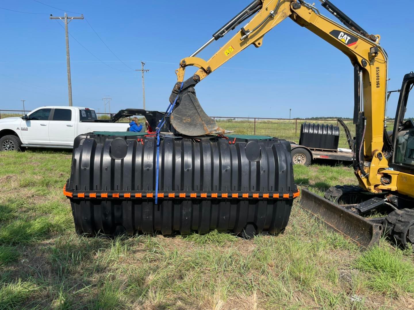 A small excavator lifts a large black septic tank with blue straps in a field. A white truck is parked nearby.