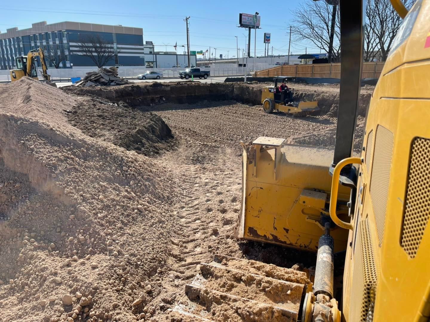 Construction site with heavy machinery: a bulldozer, excavator, and compactor, working on a large dirt pile.