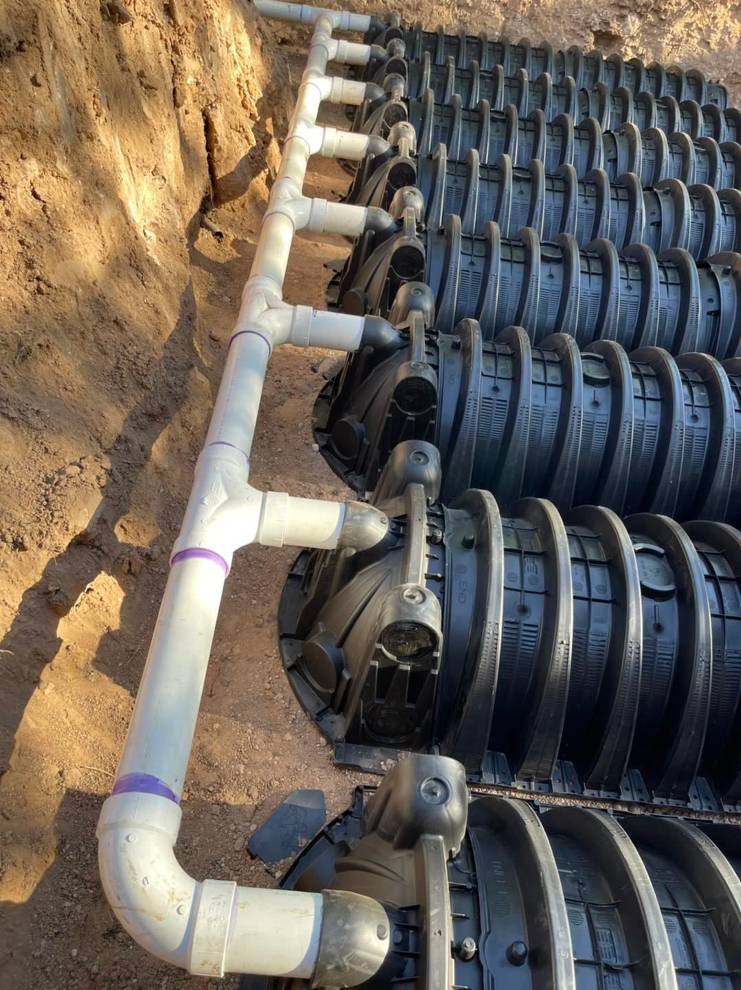 Rows of black septic chambers with white PVC piping in a dug trench.