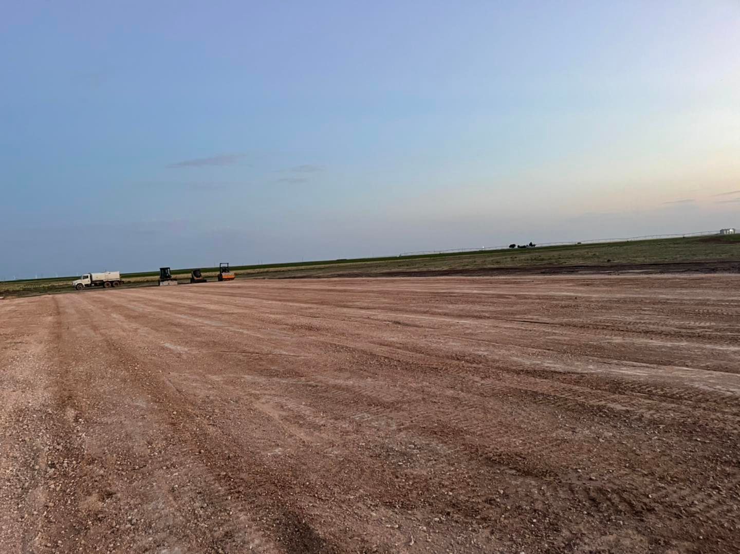 An empty, brown dirt lot with faint tractor tracks, under a pale blue sky.