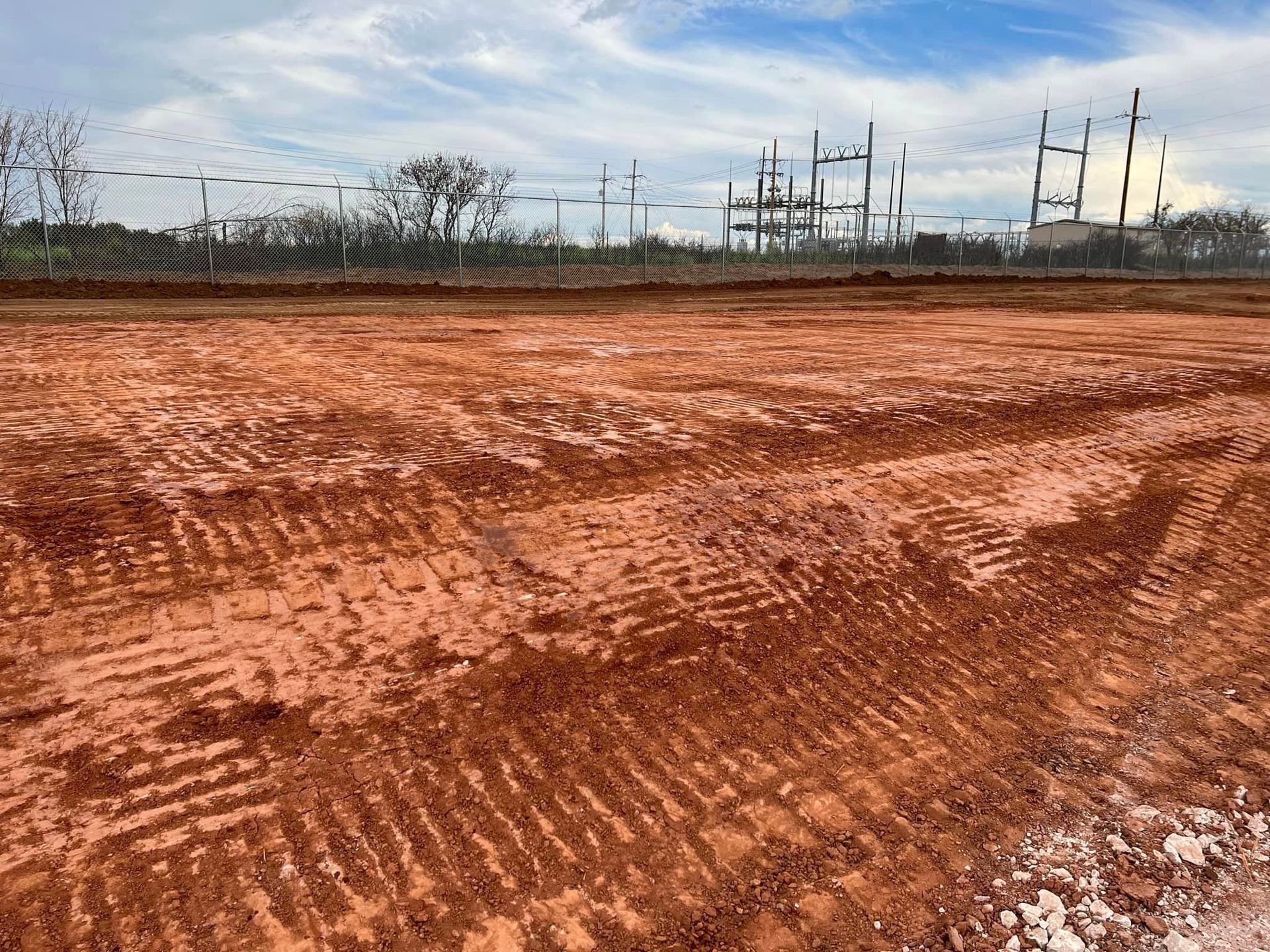 Red dirt field with tire tracks, fence, and power lines under a cloudy sky.