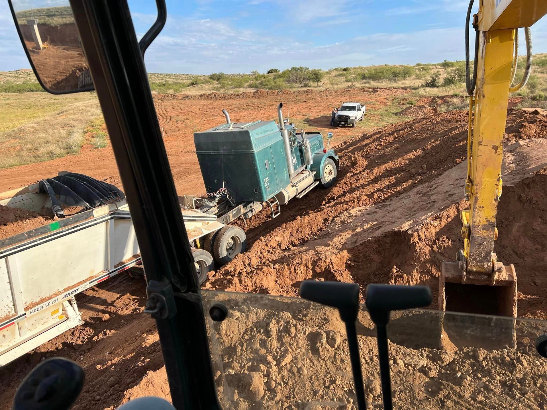 Excavator working near a semi-truck and trailer in a muddy, brown field. A white pickup truck is parked nearby.