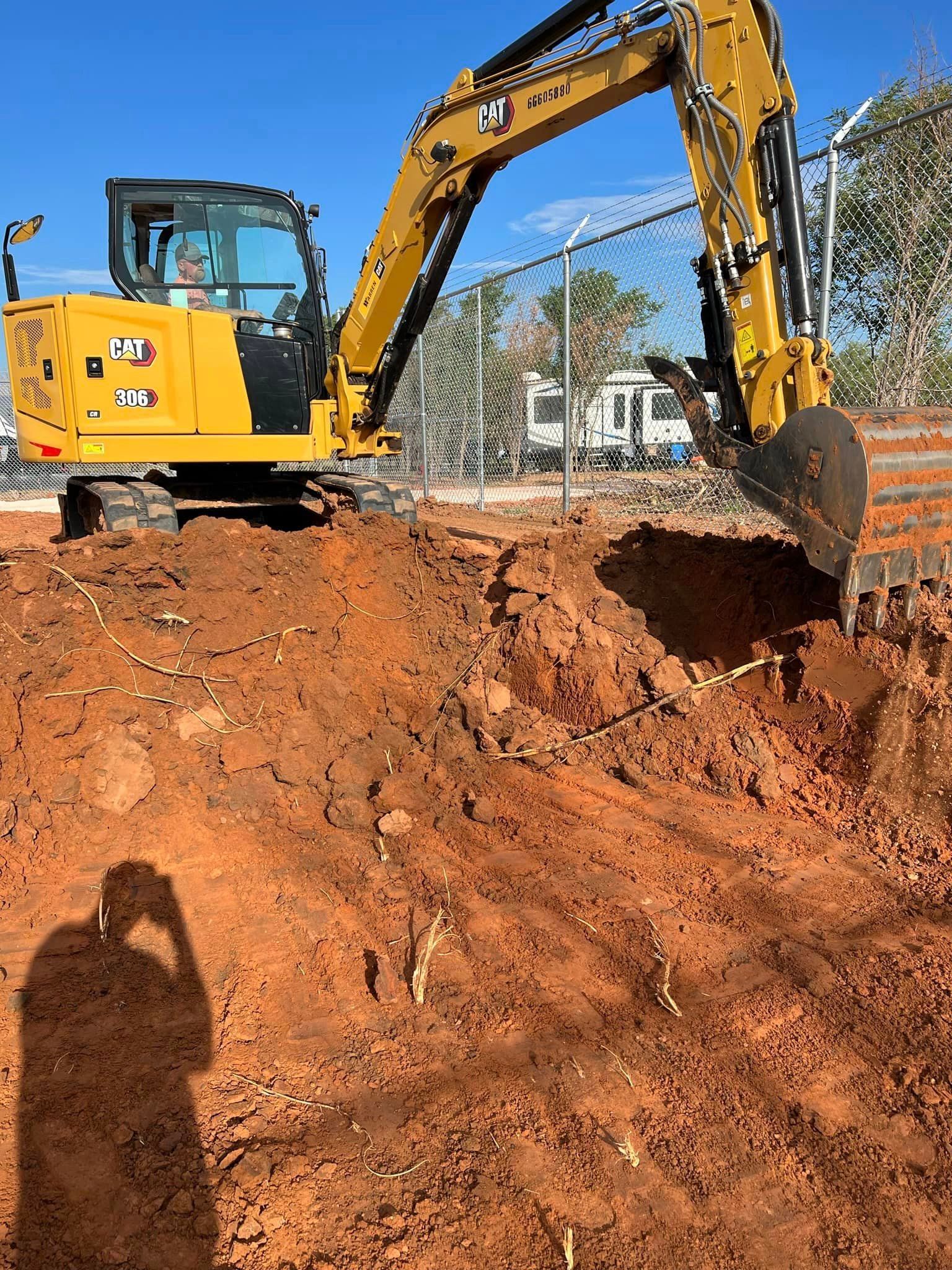 Yellow excavator digging in red dirt near a fence and buildings.