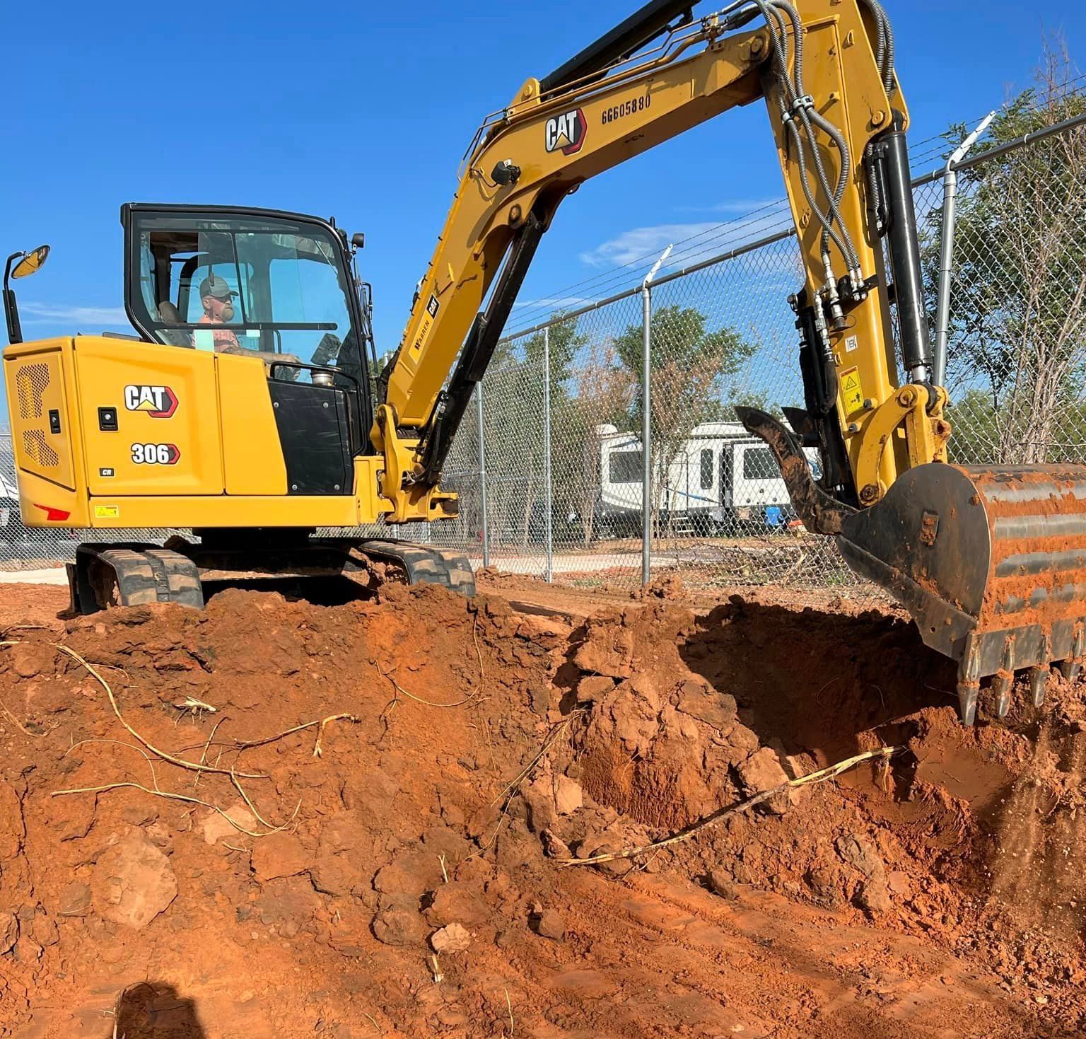 Yellow excavator digging in reddish dirt near a fence, with a clear blue sky.