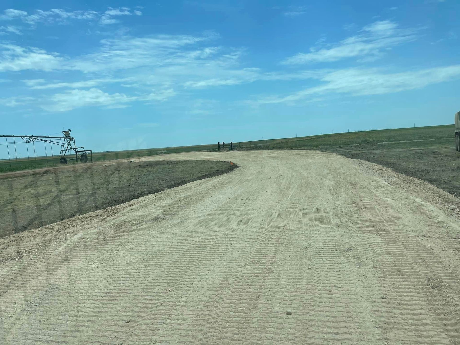 Gravel road curves upward toward a grassy hill under a blue sky, fence on the left.
