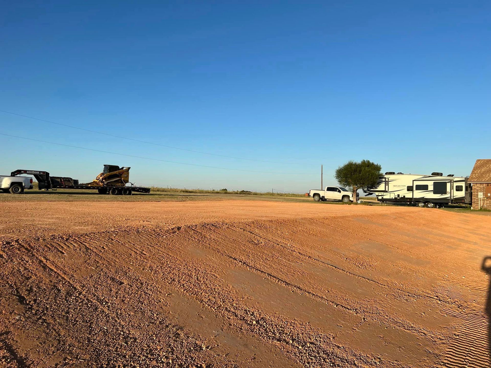 Flat, red earth with vehicle tracks. Trucks and RV on horizon under blue sky.