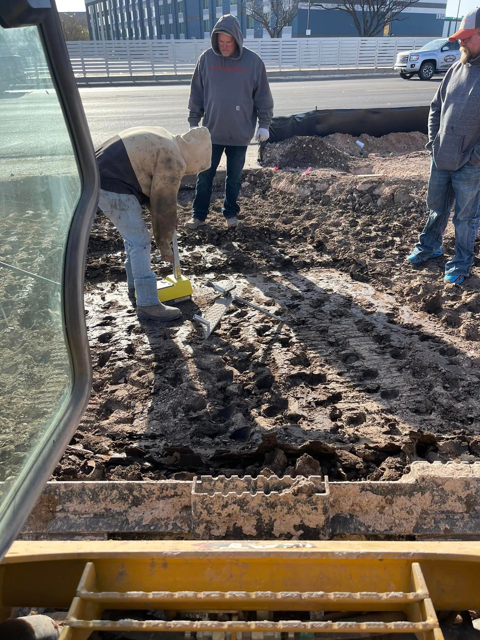 Man shovels mud from a dug-up area; two others watch in a parking lot.