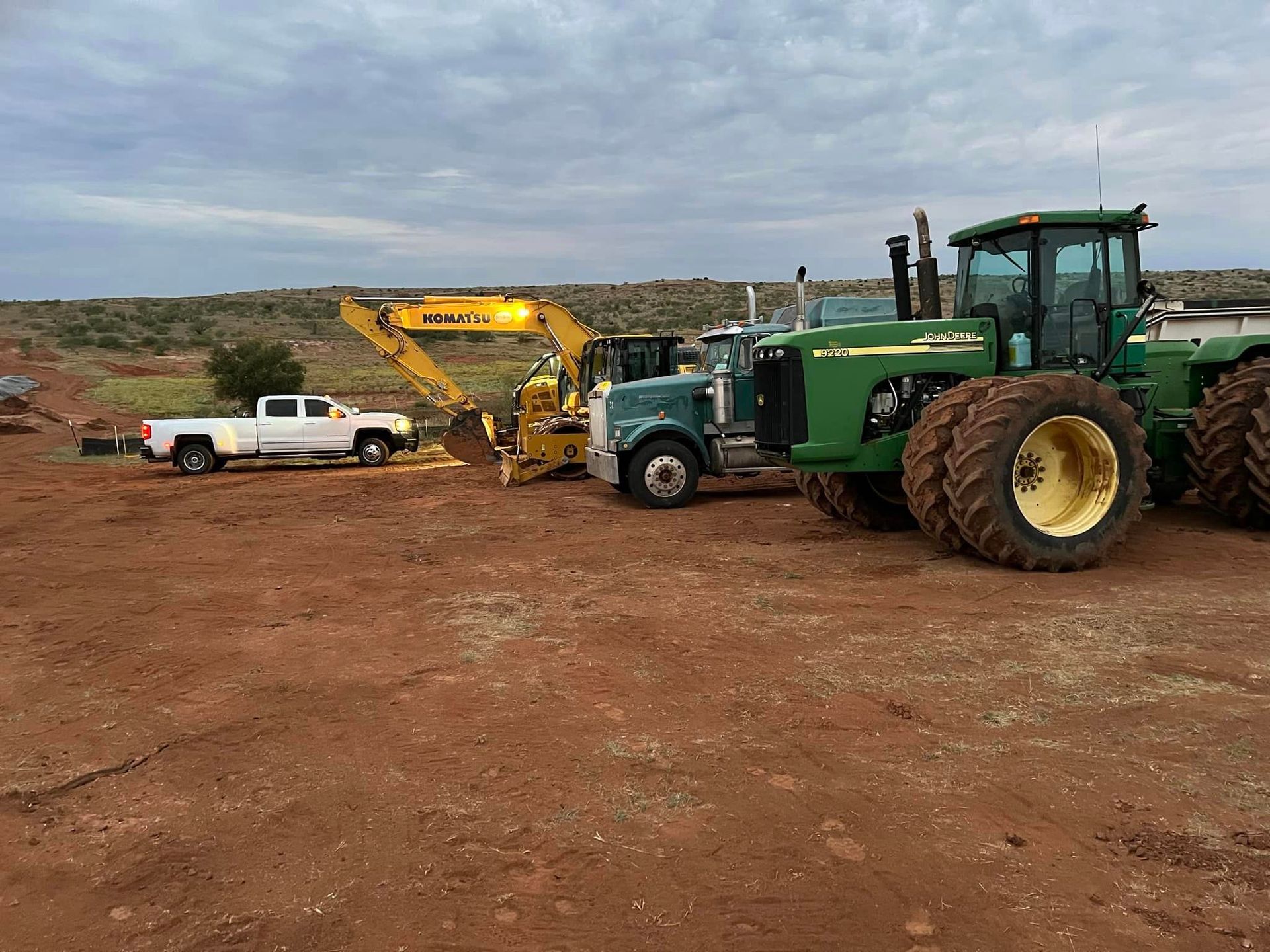 White truck, green tractor, excavator and semi-truck parked on a red dirt lot under a cloudy sky.