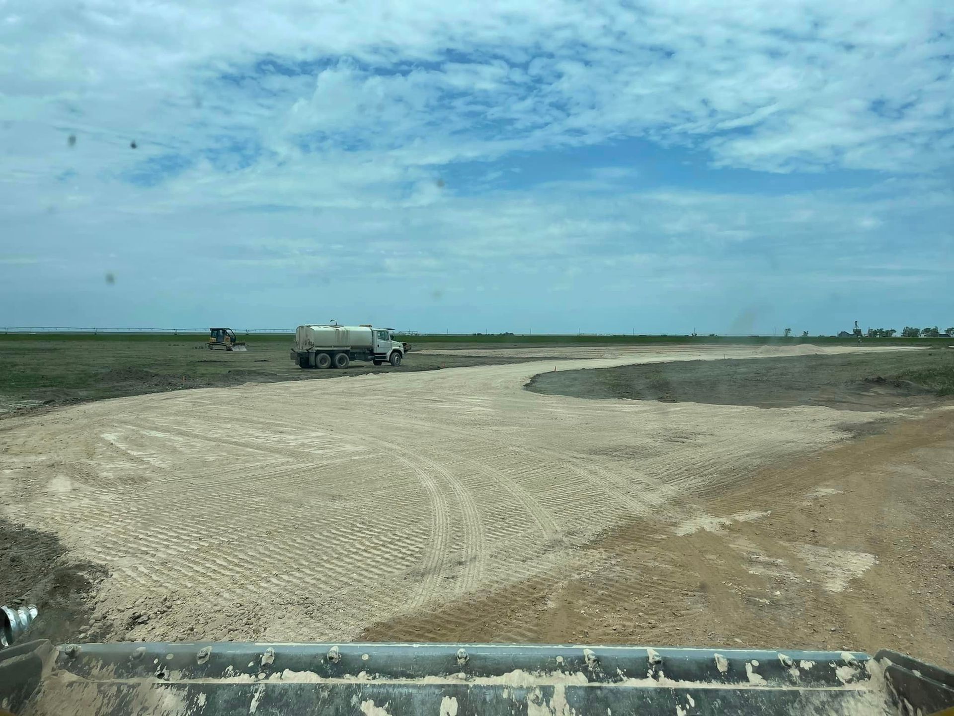 A truck sprays liquid in a field under a cloudy blue sky; construction in progress.