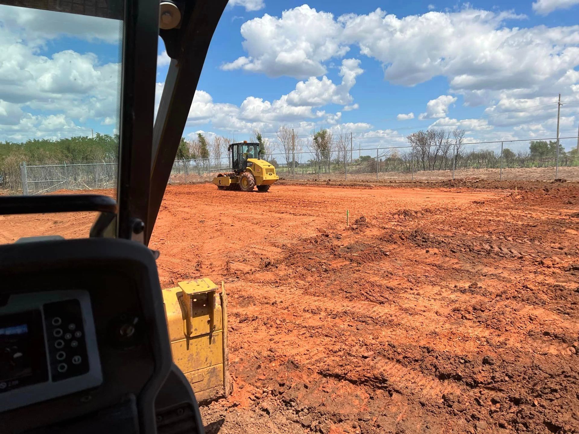 View from construction equipment cabin, looking at a roller on a red-earth construction site under a blue sky.