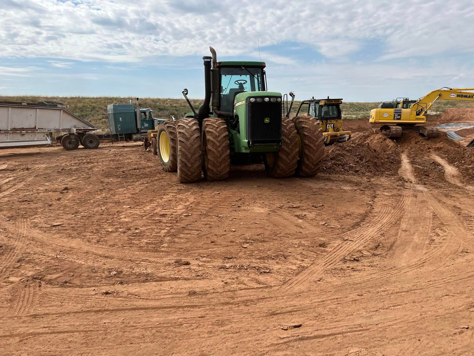 A John Deere tractor sits in a muddy field, with other construction vehicles nearby under a cloudy sky.