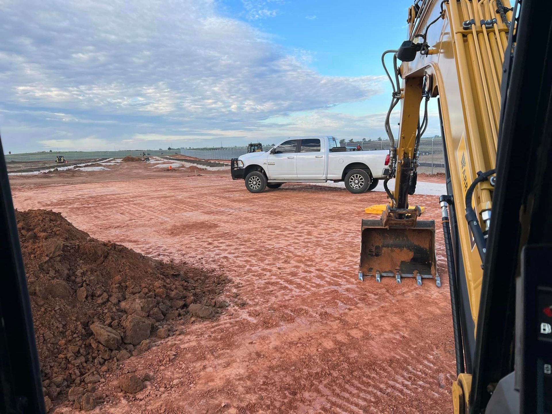 Excavator in a dirt field, white pickup truck in the background, overcast sky.