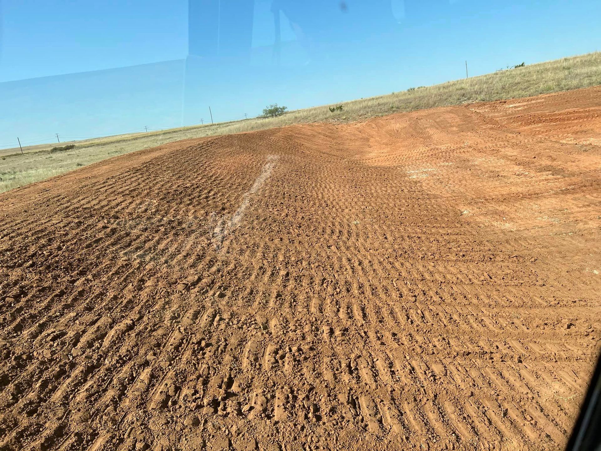 A plowed, red-brown field with tire tracks and a white line, sloping uphill towards a grassy hill under a clear sky.