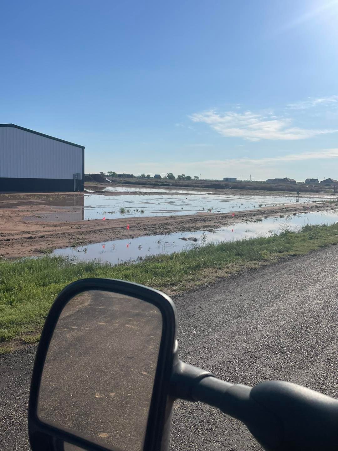 Flooded field beside a large white building, seen from a vehicle. Bright sky and asphalt road in foreground.
